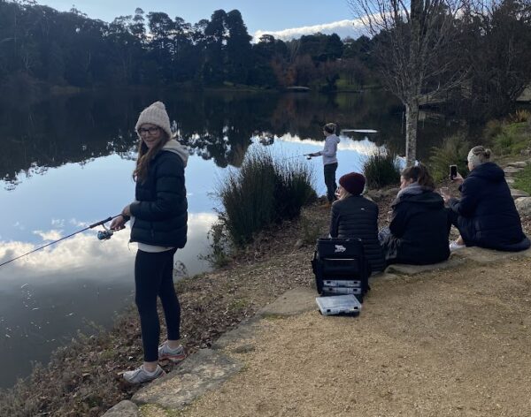 One woman is facing the camera smiling whilst fishing by a lake. Three women are looking towards the lake, with another woman fishing in the background